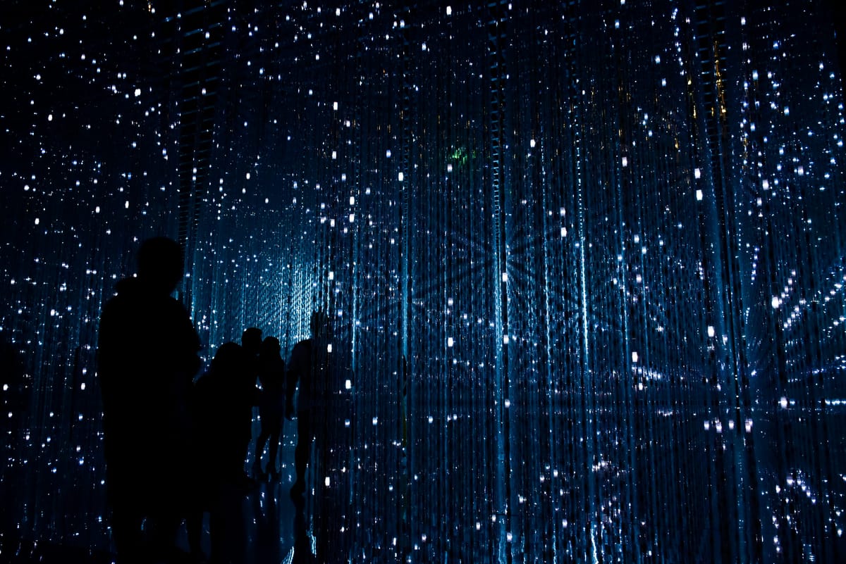 People stood in an art installation with lights dotted around the room and reflected in walls. It resembles a quantum network visualisation.